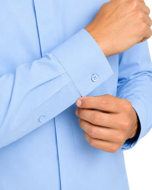 Close-up of a man wearing a light blue clerical shirt and buttoning the cuff, with visible hands and part of the torso against a plain light background.