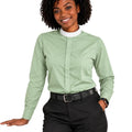 A woman modeling a sage green long-sleeve clerical shirt with a white mock neck collar, paired with black pants and a black belt, against a plain white background.