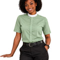 A woman modeling a sage green short-sleeve clerical shirt with a white mock neck collar, paired with black pants and a black belt, against a plain white background.