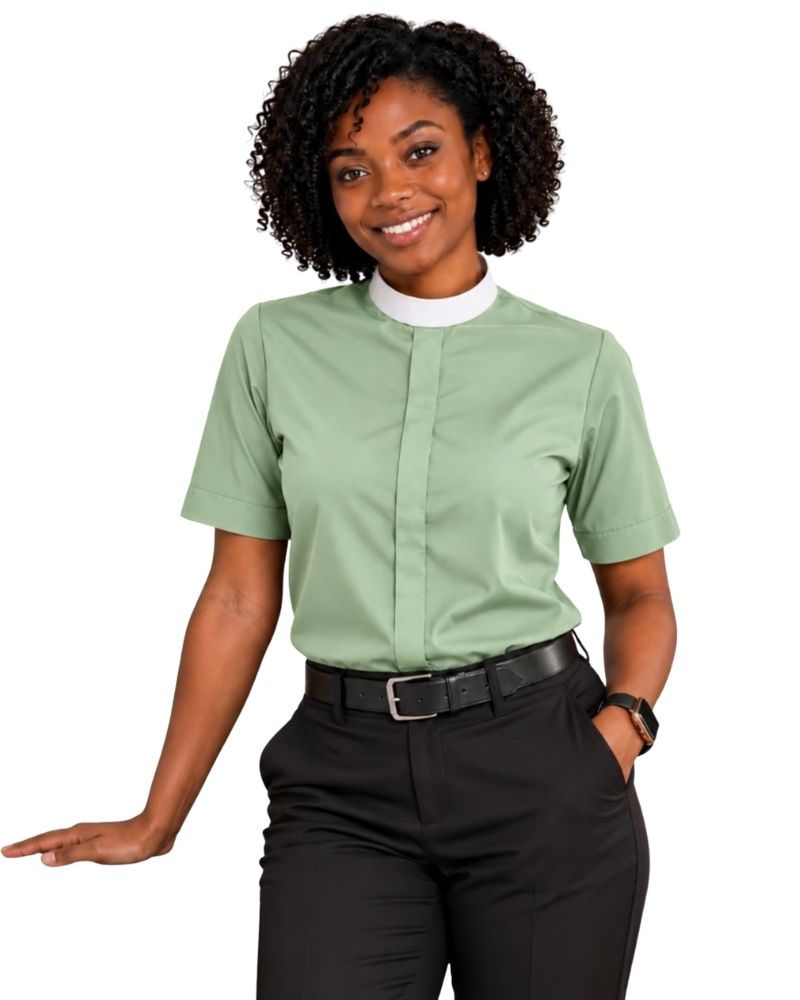 A woman modeling a sage green short-sleeve clerical shirt with a white mock neck collar, paired with black pants and a black belt, against a plain white background.