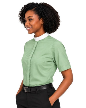 A woman modeling a sage green short-sleeve clerical shirt with a white mock neck collar, paired with black pants and a black belt, against a plain white background.