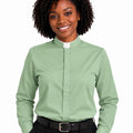 A smiling woman with curly black hair wearing a sage green long-sleeve clerical shirt with a white collar tab, paired with black trousers and a black belt, against a plain white background.