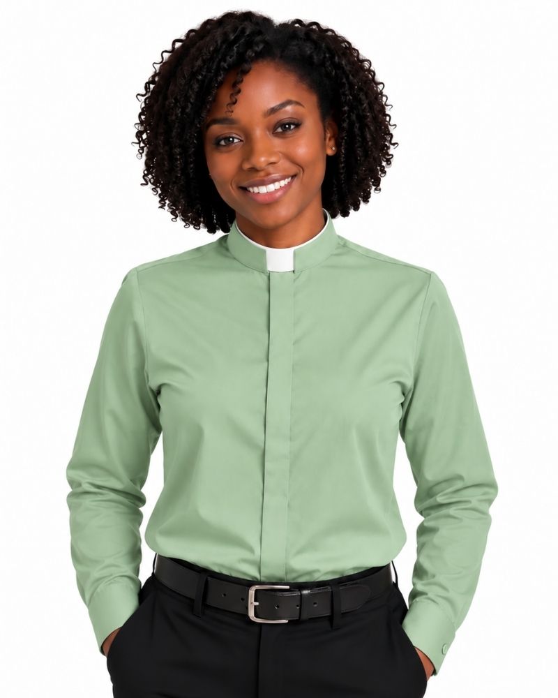 A smiling woman with curly black hair wearing a sage green long-sleeve clerical shirt with a white collar tab, paired with black trousers and a black belt, against a plain white background.