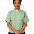 A woman wearing a sage green short-sleeve clerical shirt with a white collar, paired with black trousers, standing against a plain white background.
