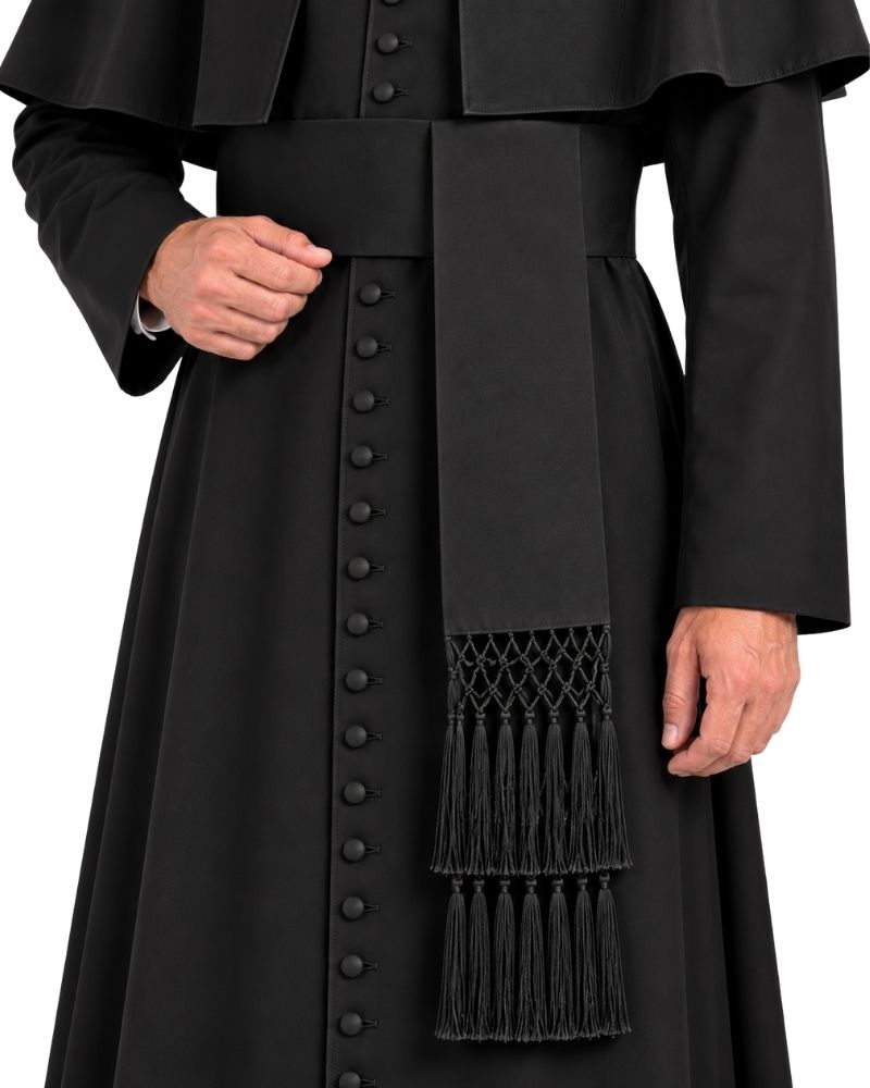 A close-up of a black clerical vestment with a wide front panel of decorative buttons, a waist sash, and a hanging embroidered tassel fringe, shown on a person against a plain white background.