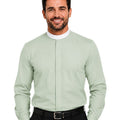 A smiling man wearing a light sage green long-sleeve clerical shirt with a white collar, paired with black trousers and a black belt, against a plain white background.