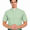 A man wearing a light sage green short-sleeve priest shirt with a white collar, paired with a black belt and black trousers, against a plain white background.