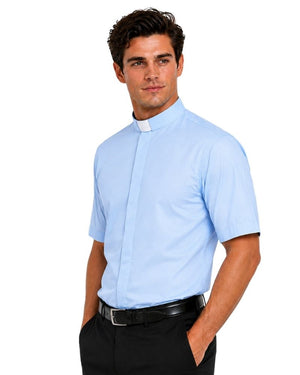 Left side view of a man wearing a light blue short sleeve clergy shirt with a white tab collar, paired with black dress pants, shown on a professional studio white background.