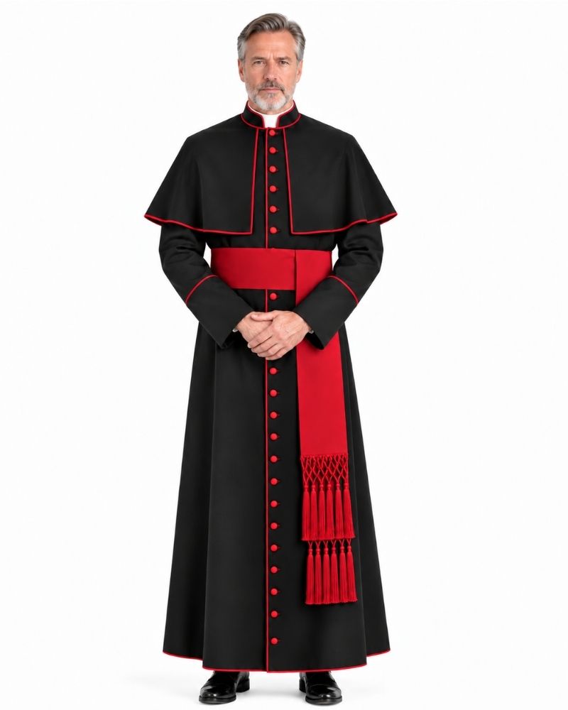 A man modeling a Traditional Clerical Cassock with Red Trim against a plain white background. The cassock features a long black robe with red piping, a wide red waist sash, and a matching red dalmatic with red tassels.