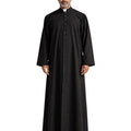 A man wearing a long black clerical robe with a high collar and a row of front buttons, standing against a plain white background.