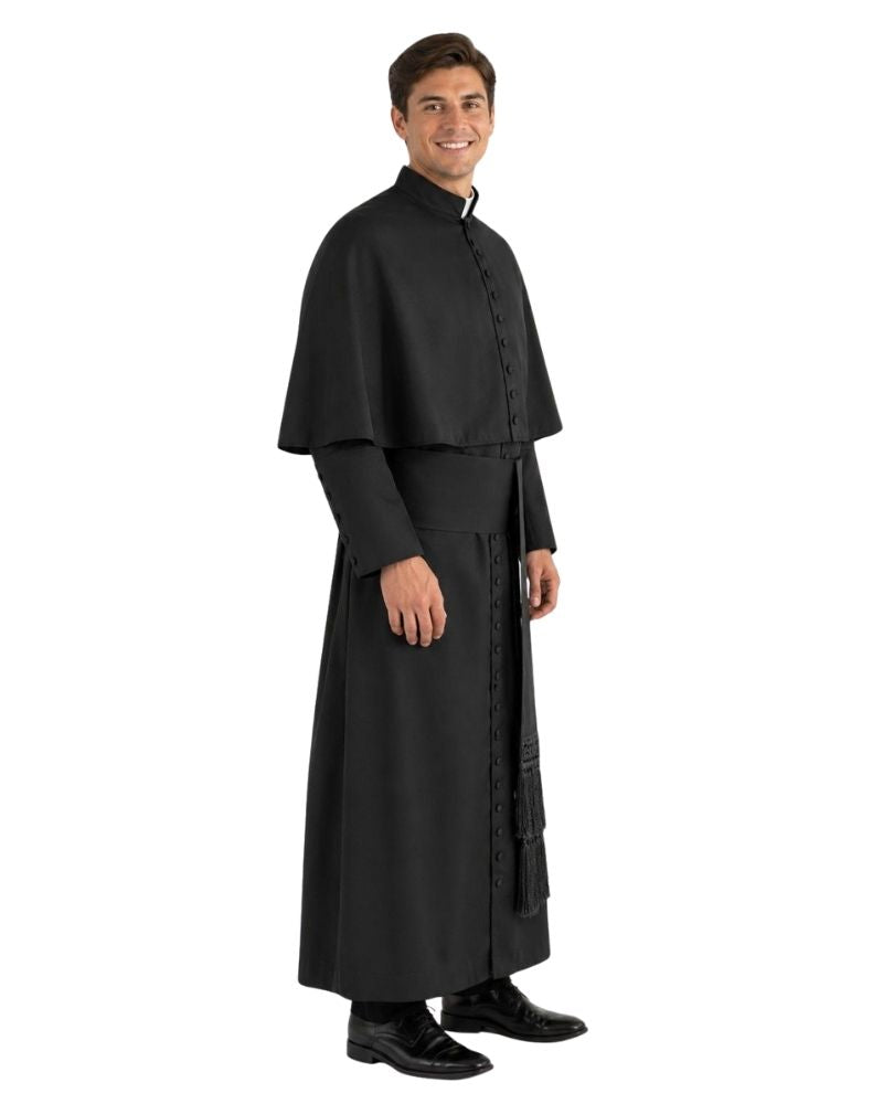 A man modeling a black priestly  consisting of a long black robe with a high collar, black cape, and black shoes, photographed against a solid white background.
