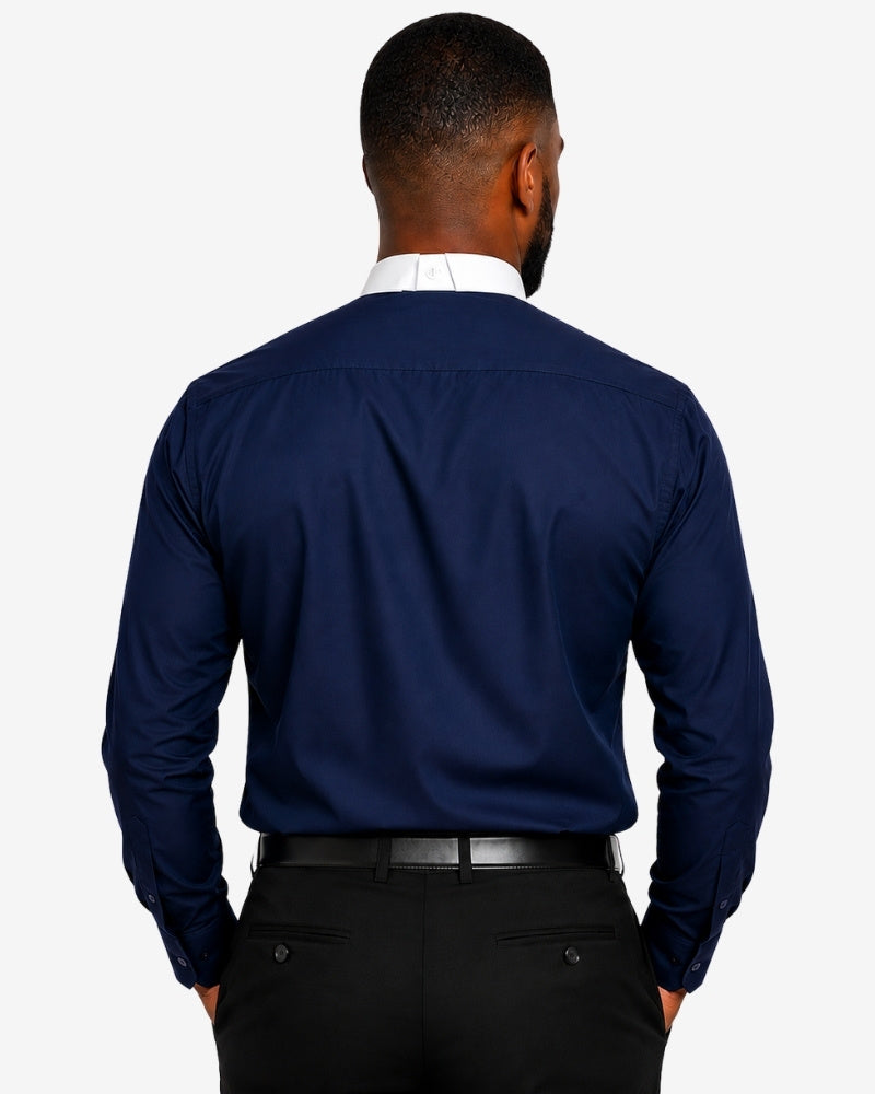 A man viewed from the back wearing a navy blue long-sleeve clerical shirt, a white collared undershirt, and black trousers against a light gray studio background.
