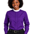 A smiling woman with curly black hair wearing a purple long-sleeve clerical shirt with a white mock neck collar, paired with black trousers and a black belt, against a plain white background.