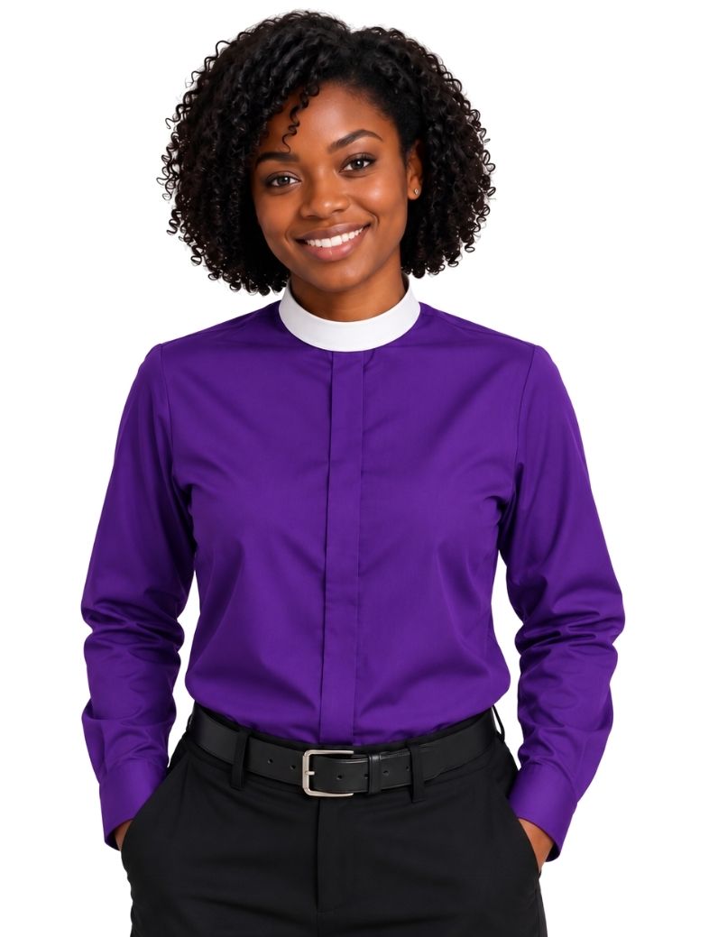 A smiling woman with curly black hair wearing a purple long-sleeve clerical shirt with a white mock neck collar, paired with black trousers and a black belt, against a plain white background.