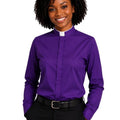 A smiling woman with curly black hair wearing a purple long-sleeve clerical shirt with a white collar, paired with black trousers and a black belt, against a plain white background.