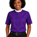 A smiling woman with curly black hair wearing a purple short-sleeve clerical shirt with a white mock neck collar, paired with black trousers and a black belt, against a plain white background.