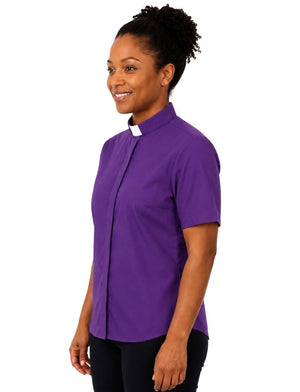 A woman wearing a purple short-sleeve clerical shirt with a white clerical collar, paired with black pants, photographed against a plain white background.