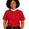 A smiling woman with curly black hair wearing a bright red short-sleeve clerical shirt with a white mock neck collar, paired with black trousers and a black belt, against a plain white background.