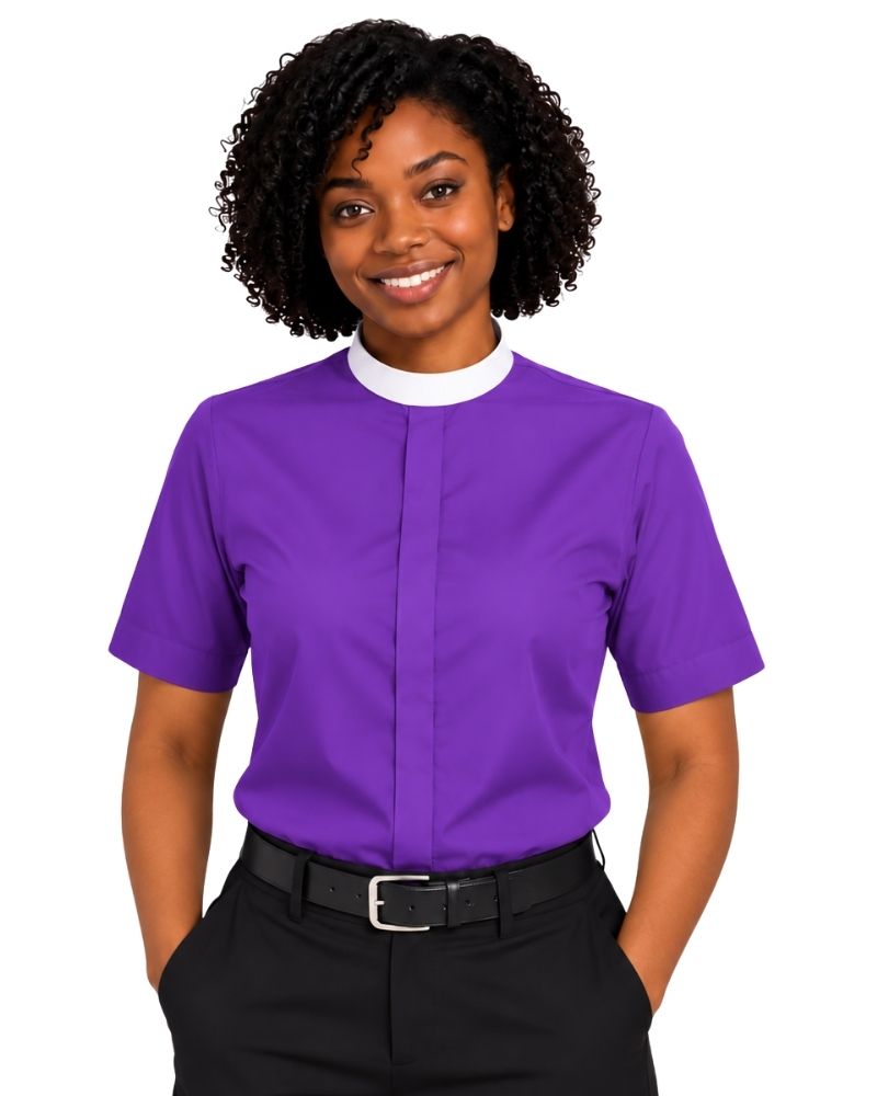 A woman with curly black hair wearing a purple short-sleeve clerical shirt with a white mock neck collar, paired with black trousers and a black belt, against a plain white background.