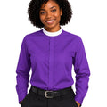 A woman with curly black hair wearing a vibrant purple long-sleeve clerical shirt with a contrasting white mock neck collar, paired with black trousers and a black belt, against a plain white background.