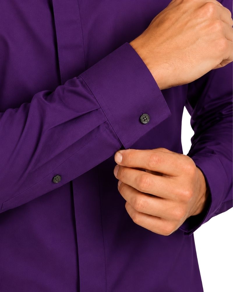 Close-up of a man wearing a solid purple long-sleeve clerical shirt and buttoning the cuff, with the hands and torso visible against a plain white background.