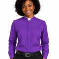 A smiling woman with curly black hair wearing a purple long-sleeve clerical shirt with a white collar, paired with black trousers and a black belt, against a plain white background.