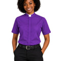 A smiling woman with curly black hair wearing a purple short-sleeve clerical shirt with a white collar tab, paired with black trousers and a black belt, against a plain white background.