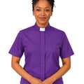 A woman wearing a purple short-sleeve clerical shirt with a white collar, standing against a plain white background.