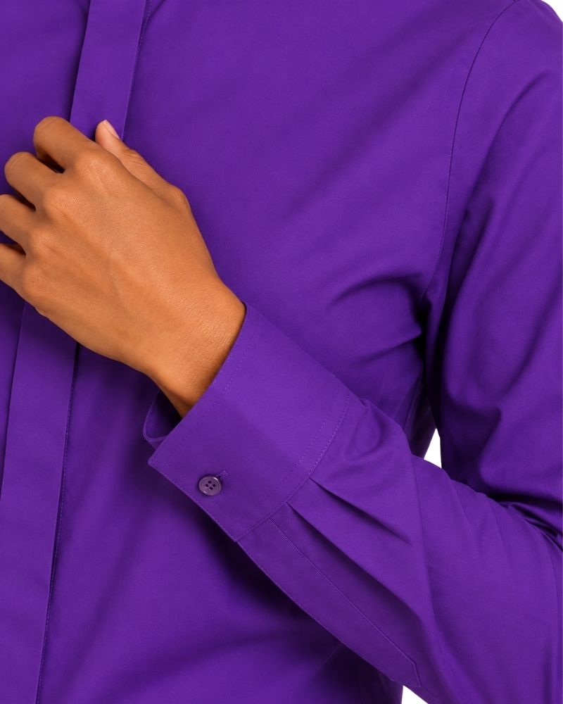 A close-up of a woman wearing a long-sleeve clerical shirt in a vivid purple color, showing the hand adjusting the collar and the cuff with a button closure.
