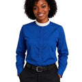 A woman with curly black hair wearing a royal blue long-sleeve clerical shirt with a white mock neck collar, paired with black trousers and a black belt, against a plain white background.