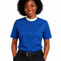 A smiling woman with curly black hair wearing a royal blue short-sleeve clerical shirt with a white mock neck trim, paired with black trousers and a black belt, against a plain white background.