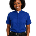 A smiling woman with curly black hair wearing a royal blue short-sleeve clerical shirt with a white collar, paired with black trousers and a black belt, against a plain white background.