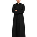 A man modeling a long black clerical robe with a white collar, standing with arms crossed against a plain white background.