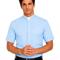 A man wearing a light blue short-sleeve clerical shirt with a white collar, paired with black trousers and a black belt, against a plain white background.
