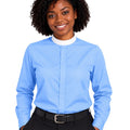 A smiling woman with curly black hair wearing a light blue long-sleeve clerical shirt with a white mock neck collar, paired with black trousers and a black belt, against a plain white background.