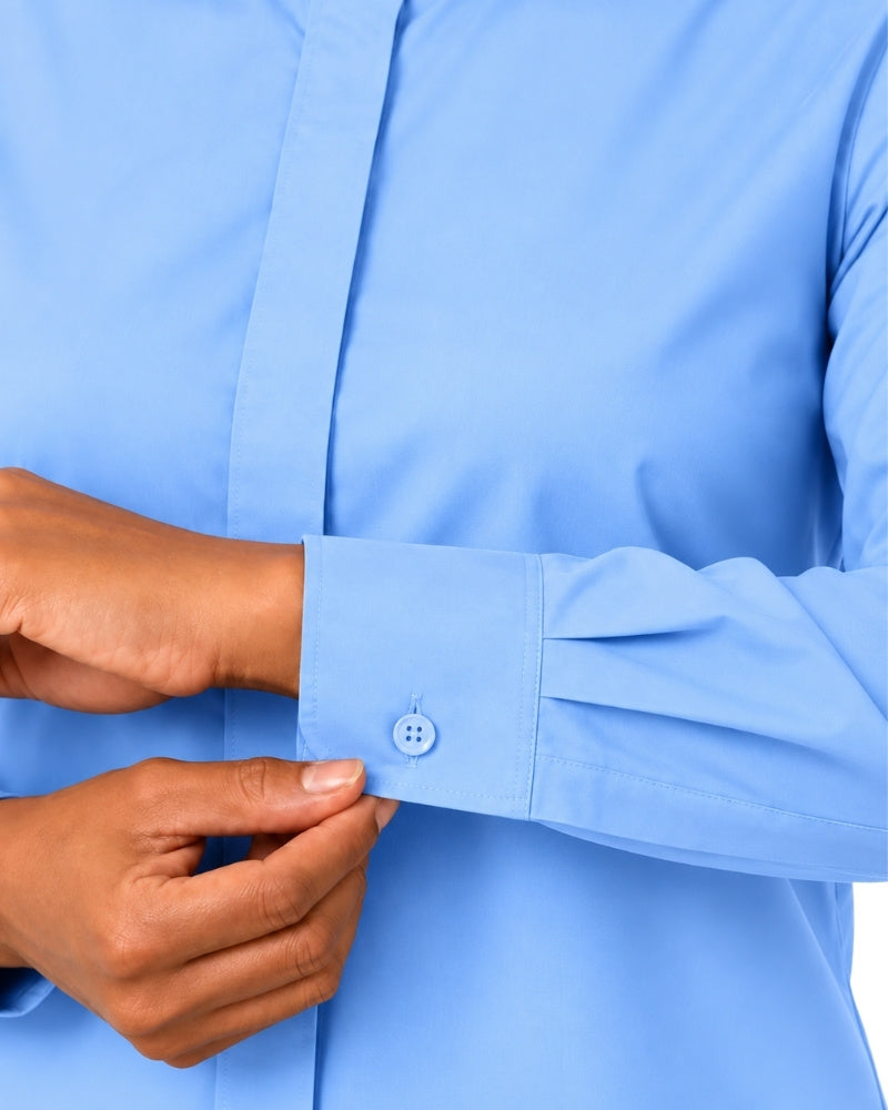 A close-up of a woman hands adjusting the cuff of a light blue long-sleeve clerical shirt, showing the buttoned cuff and fabric texture.