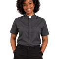 A smiling woman with curly black hair wearing a dark gray short-sleeve clerical shirt with a white collar and black trousers, standing against a plain white background.