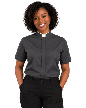 A smiling woman with curly black hair wearing a dark gray short-sleeve clerical shirt with a white collar and black trousers, standing against a plain white background.