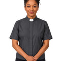 A woman wearing a dark gray short-sleeve clerical shirt with a white collar, standing against a plain white background.