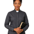 A woman wearing a dark gray long-sleeve clerical shirt with a white collar, holding a black book against a plain white background.