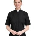 A woman wearing a black short-sleeve clerical shirt with a white collar, standing against a plain white background.