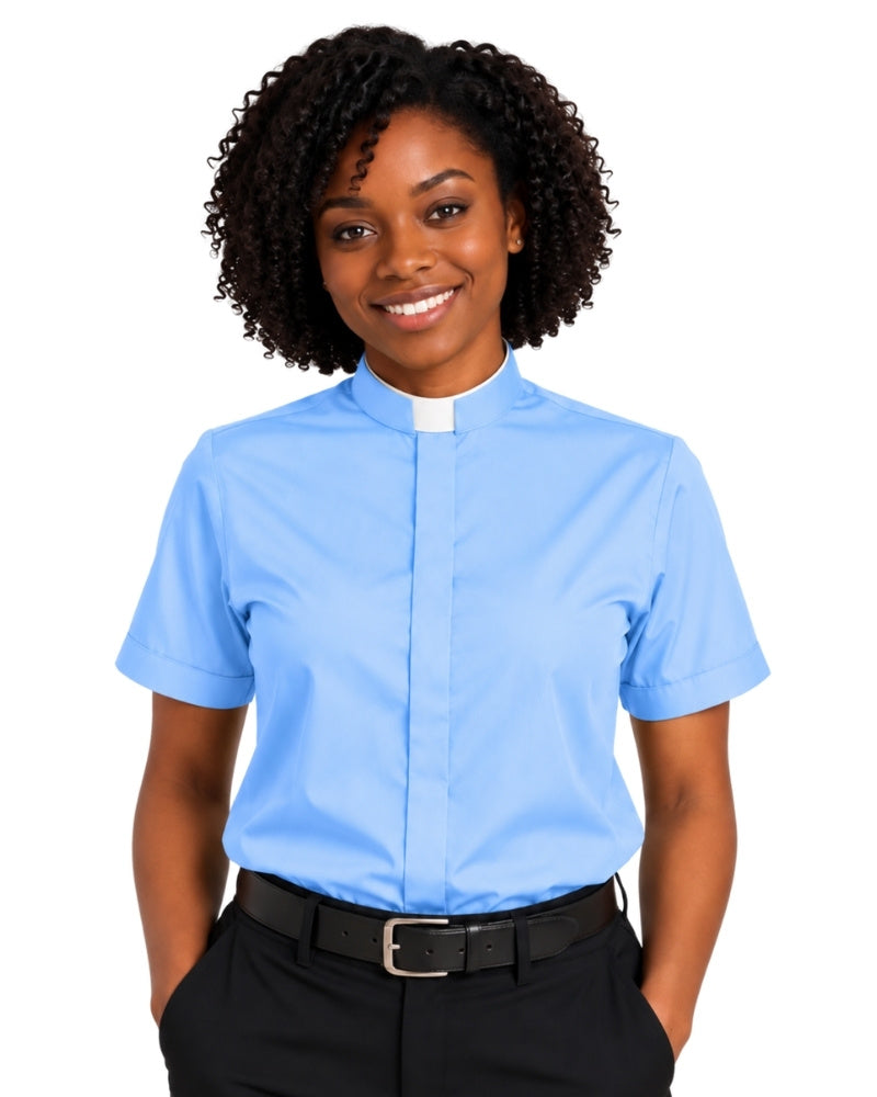 A smiling woman with curly black hair wearing a light blue short-sleeve clerical shirt with a white collar tab, paired with black trousers and a black belt, against a plain white background.