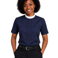 A smiling woman with curly black hair wearing a navy blue short-sleeve clerical shirt with a white mock neck collar, paired with black trousers and a black belt, against a plain white background.