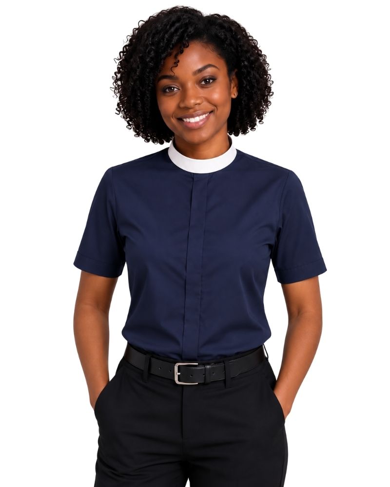 A smiling woman with curly black hair wearing a navy blue short-sleeve clerical shirt with a white mock neck collar, paired with black trousers and a black belt, against a plain white background.