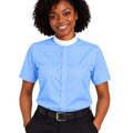 A smiling woman with curly black hair wearing a light blue short-sleeve clerical shirt with a white mock neck collar, paired with black trousers and a black belt, against a plain white background.