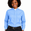 A smiling woman with curly black hair wearing a light blue long-sleeve clerical shirt with a white collar tab, paired with black trousers and a black belt, against a plain white background.