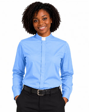 A smiling woman with curly black hair wearing a light blue long-sleeve clerical shirt with a white collar tab, paired with black trousers and a black belt, against a plain white background.