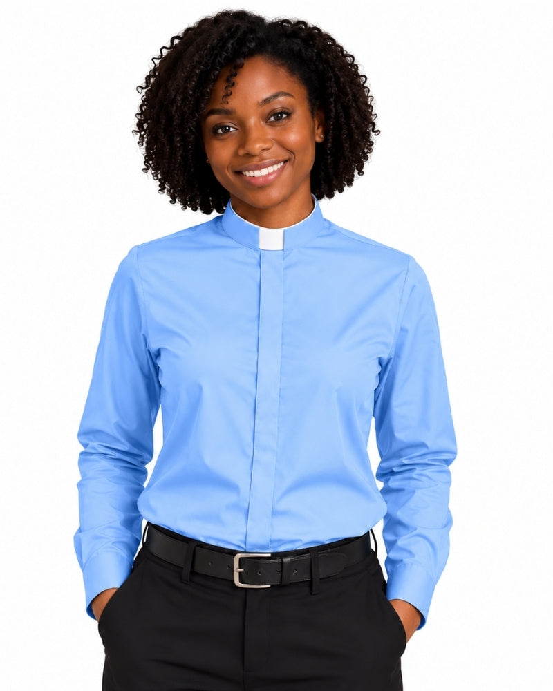 A smiling woman with curly black hair wearing a light blue long-sleeve clerical shirt with a white collar tab, paired with black trousers and a black belt, against a plain white background.