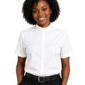 A smiling woman with curly black hair wearing a white short-sleeve clerical shirt with a high mock neck, tucked into black trousers with a black belt, against a plain white background.
