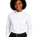 A woman with curly black hair wearing a white long-sleeve clerical shirt with a mock neck, paired with black trousers and a black belt, against a plain white background.